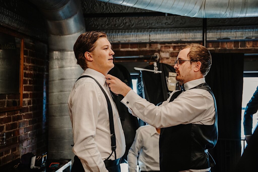A groom stands in a brick room while one of the groomsmen adjusts his bow tie as they get ready for the wedding day