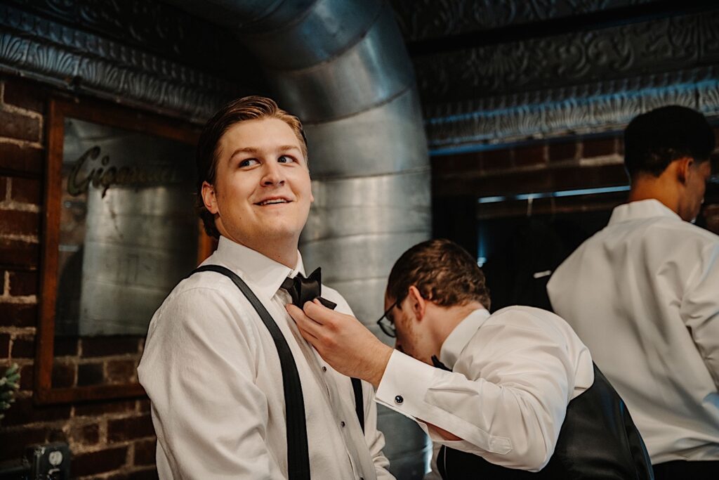 A groom smile while looking at something off camera in a brick room as a groomsmen adjusts his bow tie as they get ready for their wedding at Venue CU in Champaign