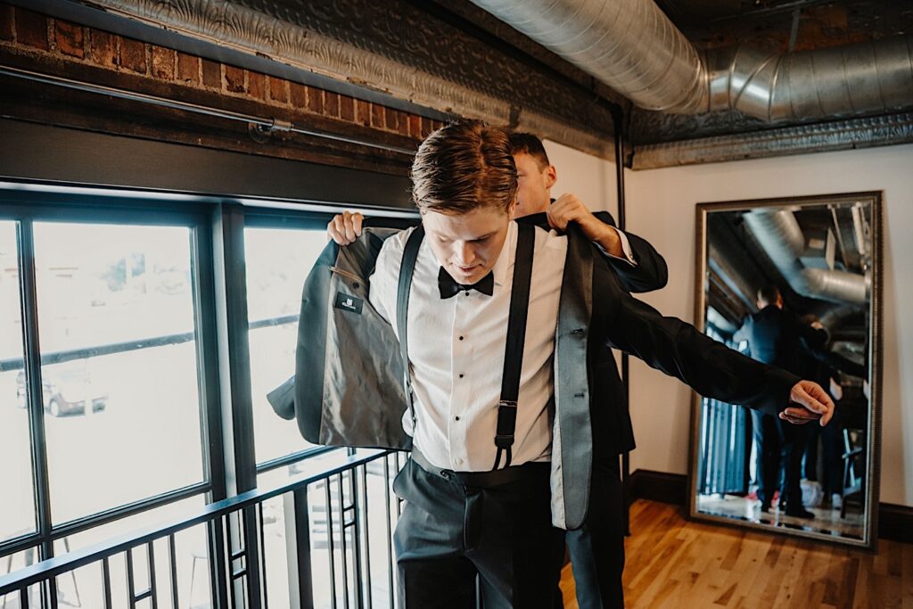 A groom puts his suit coat on with the help of his groomsmen as they get ready for his wedding day on location at Venue CU in Champaign