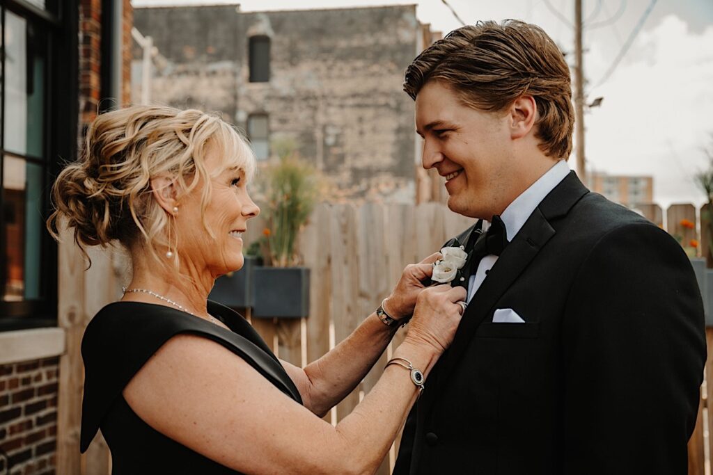 A groom smiles at his mother who smiles back at him while pinning his boutonniere on and smiling back at him while outside the wedding venue, Venue CU in Champaign