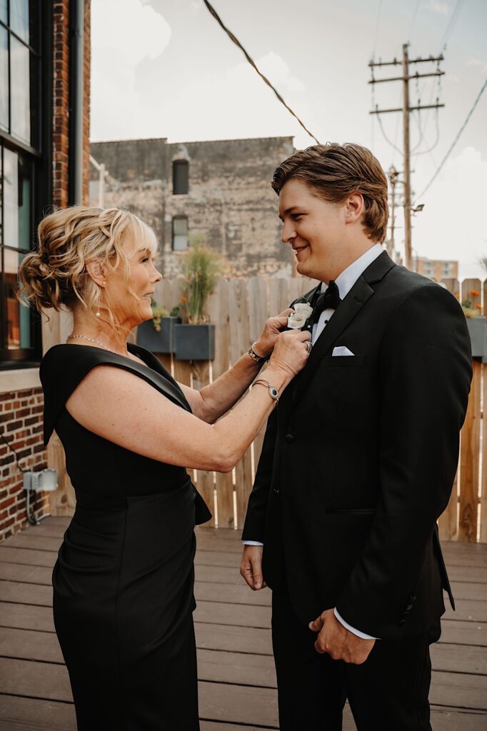 A mother smiles while pinning a boutonniere on her son as he smiles back to her before his wedding day