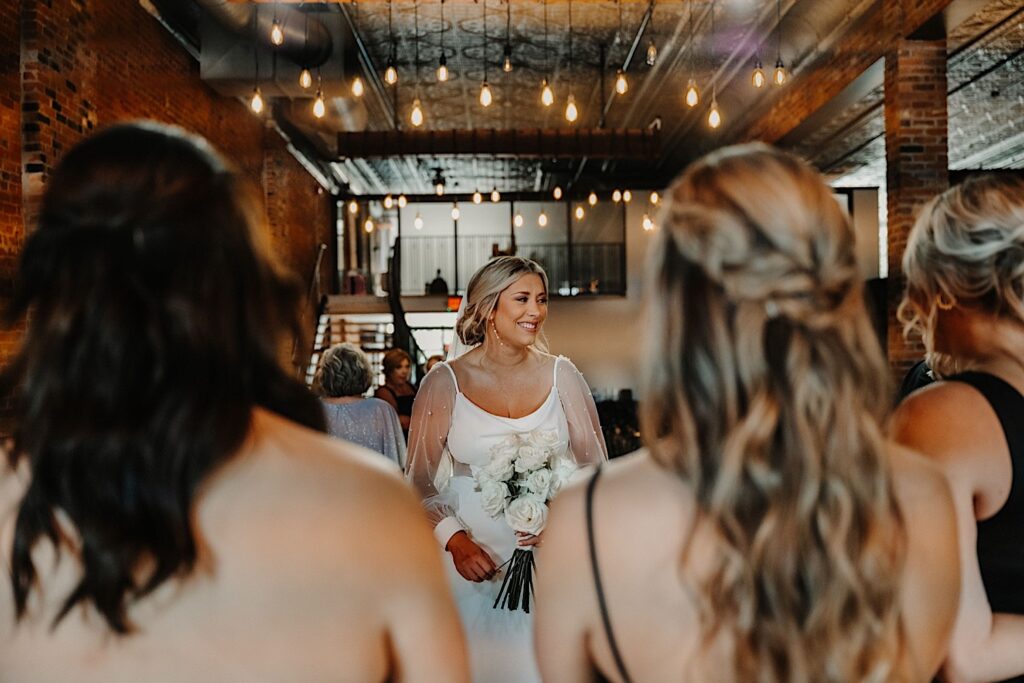 A bride smiles with her bridesmaids in the foreground of the photo facing away from the camera, this is during the bride's first look with them in her wedding venue, Venue CU in Champaign