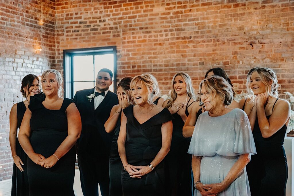 A group of bridesmaids and wedding guests smile and react seeing the bride for the first time in her wedding dress off camera while standing in their wedding venue, Venue CU in Champaign