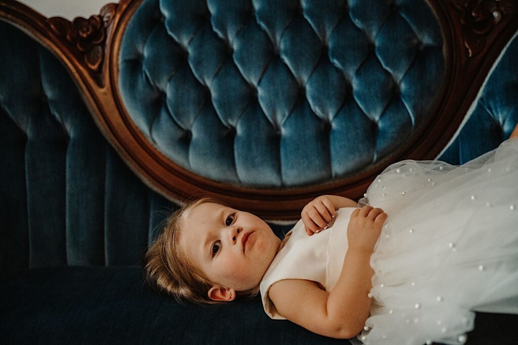 A young girl in a white dress looks at the camera while laying on a blue couch before her father's wedding