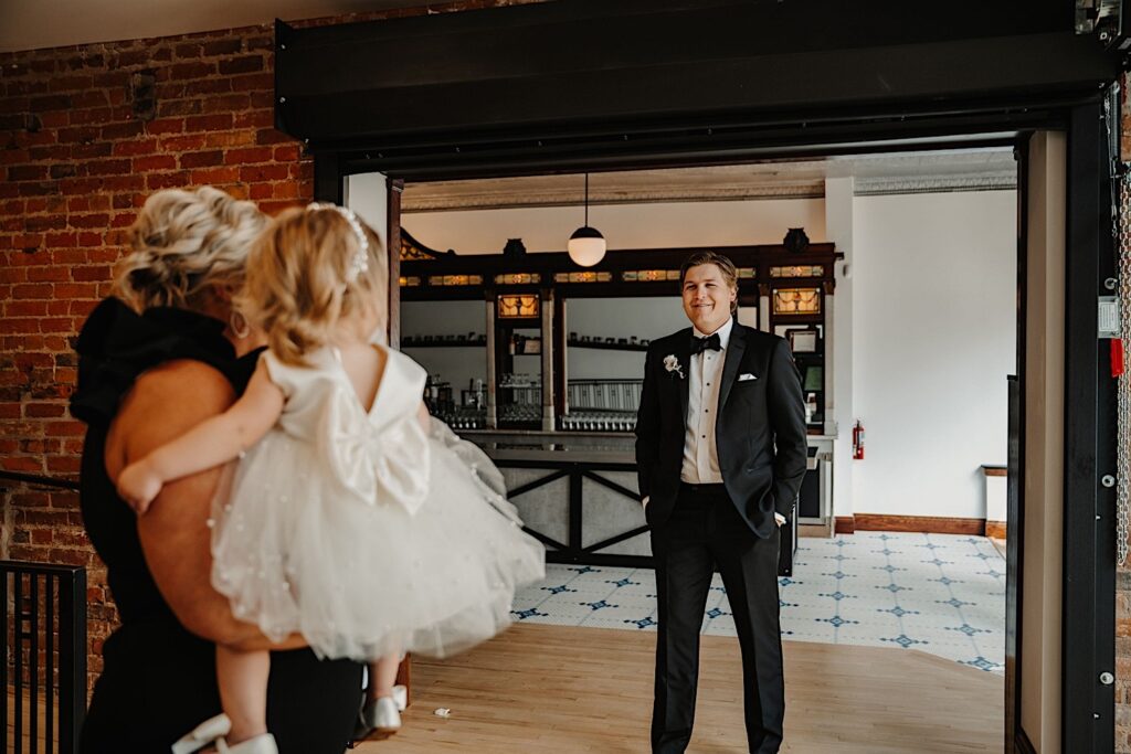 A groom smiles as his young daughter in a white dress is being carried to him by a woman