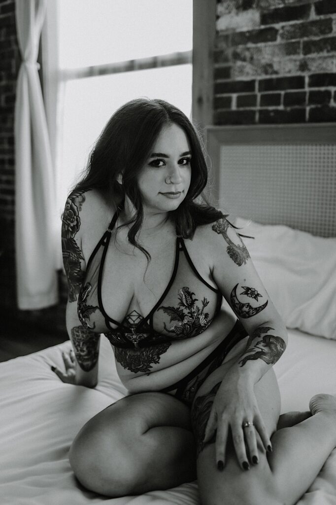 Black and white photo of a tattooed woman in lingerie sitting on a bed and looking at the camera