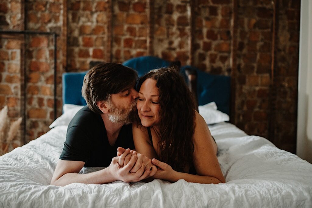 A man and woman lay in a bed facing the camera during their couples boudoir session, the man is kissing the woman on her cheek as they hold hands