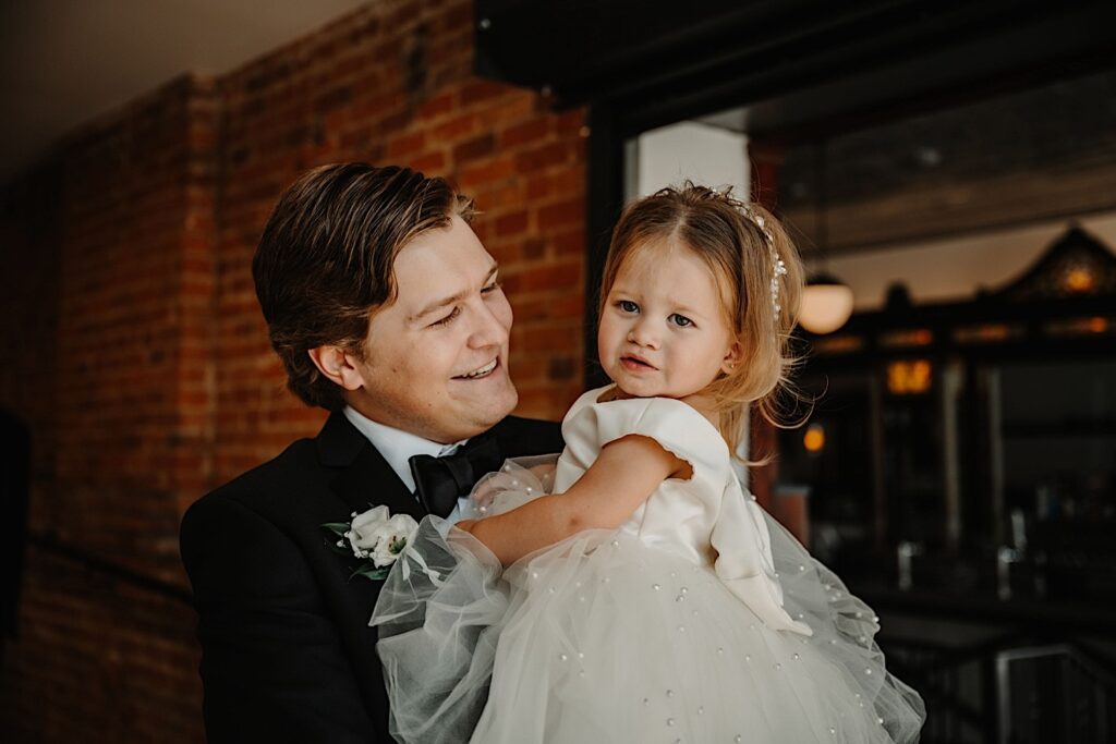 A groom smiles at his young daughter who he is holding while standing in his wedding venue, Venue CU in Champaign. The young girl is looking at the camera and is wearing a white dress