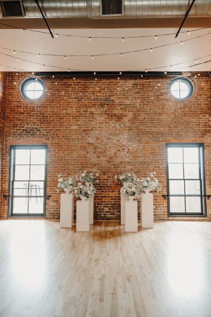 The center piece of a wedding ceremony sits in front of a brick wall before a wedding ceremony begins, the center piece consists of 6 pillars with white flowers on them