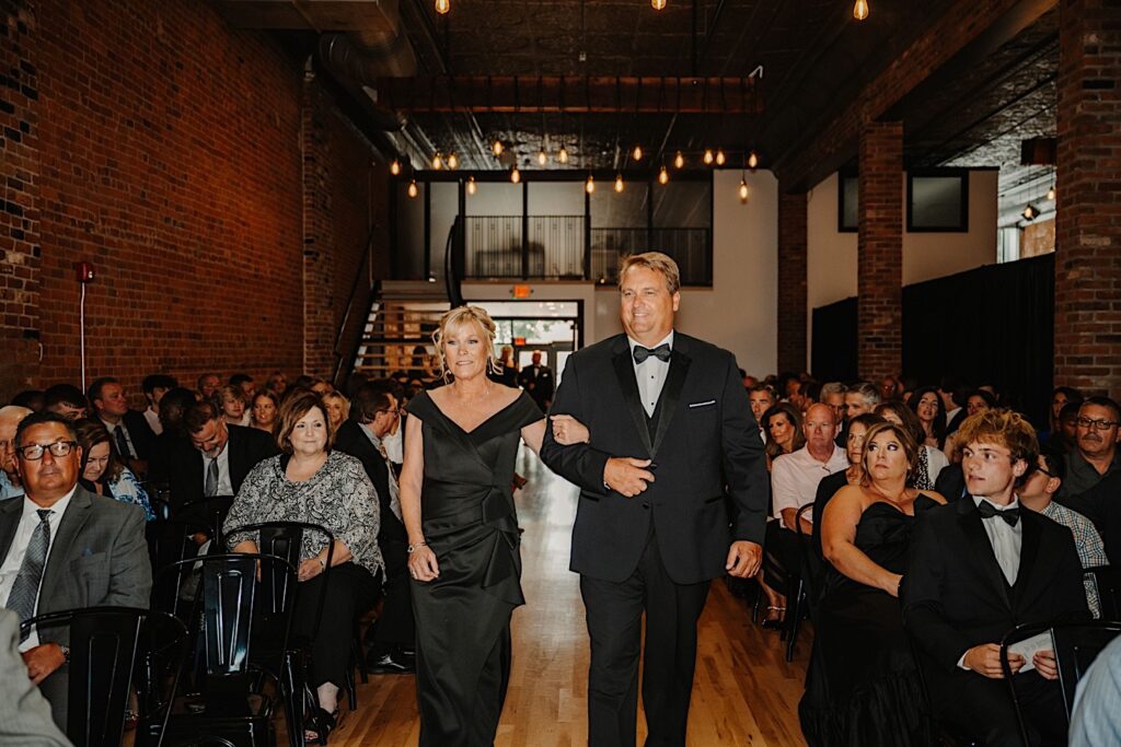 A father and mother of the groom walk down the aisle together as guests watch during an indoor wedding reception at Venue CU in Champaign