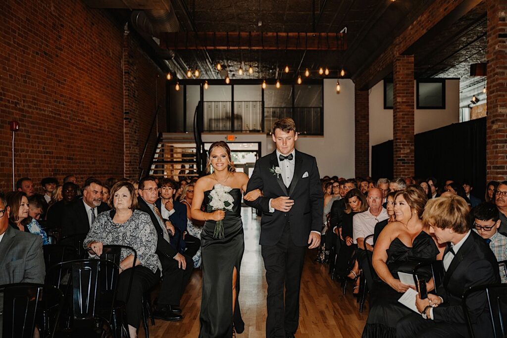 A bridesmaid and groomsman walk down the aisle together while guests watch during a wedding ceremony at Venue CU in Champaign