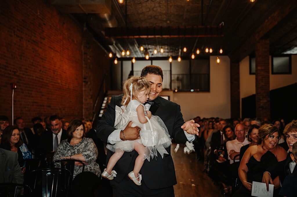 A groomsmen carries the groom's two year old daughter down the aisle of a wedding ceremony as the two toss flowers and guests watch during a wedding ceremony at Venue CU in Champaign