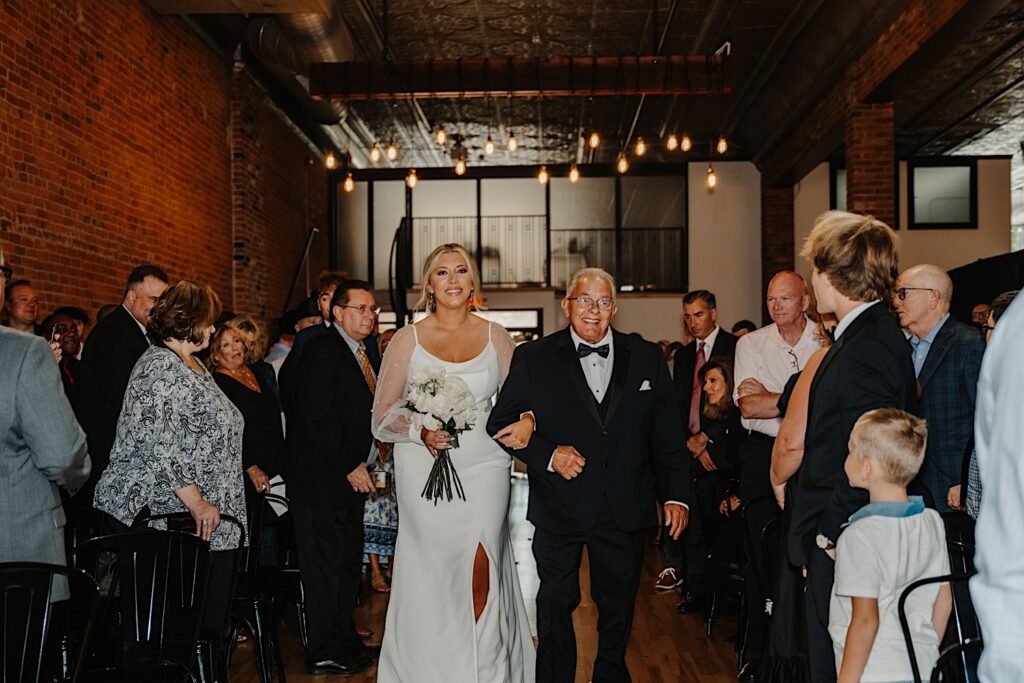 A bride smiles as while walking down the aisle of her wedding with her grandfather as guests stand and watch during a wedding ceremony at Venue CU in Champaign