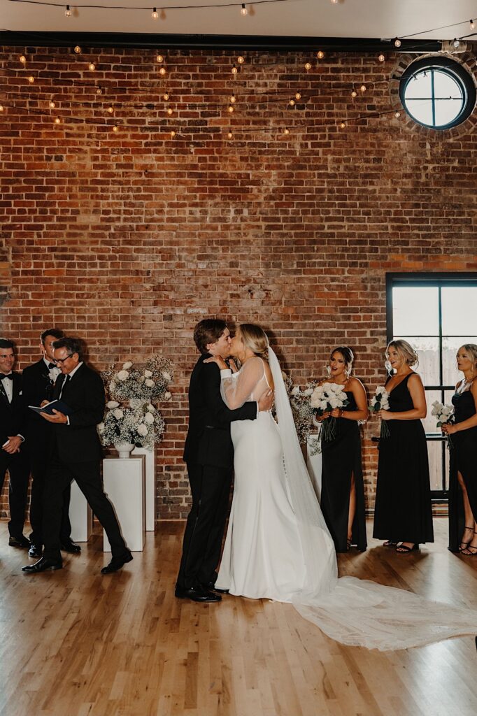 A bride and groom kiss in front of a brick wall as their wedding parties cheer during their wedding reception