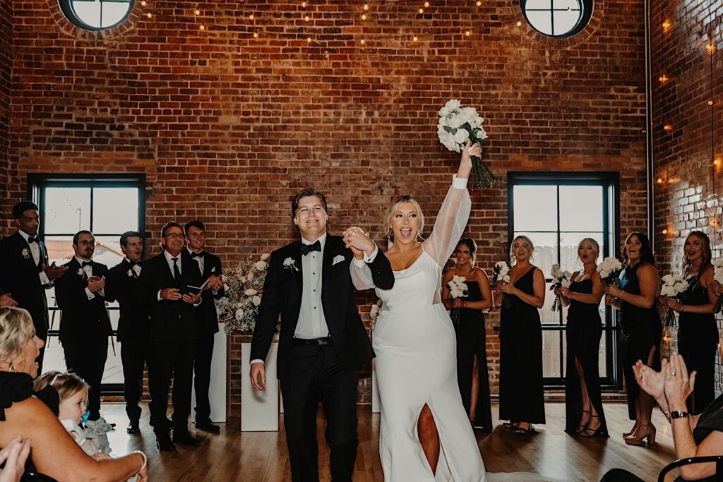 A bride and groom hold hands, smile, and exclaim while in a brick room during their indoor wedding ceremony at Venue CU in Champaign as their wedding party members cheer behind them
