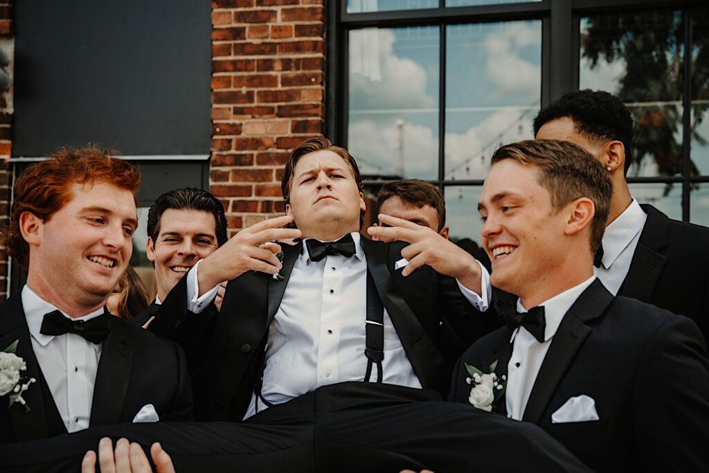 A groom is carried by his groomsmen while looking at the camera and showing piece signs to the camera