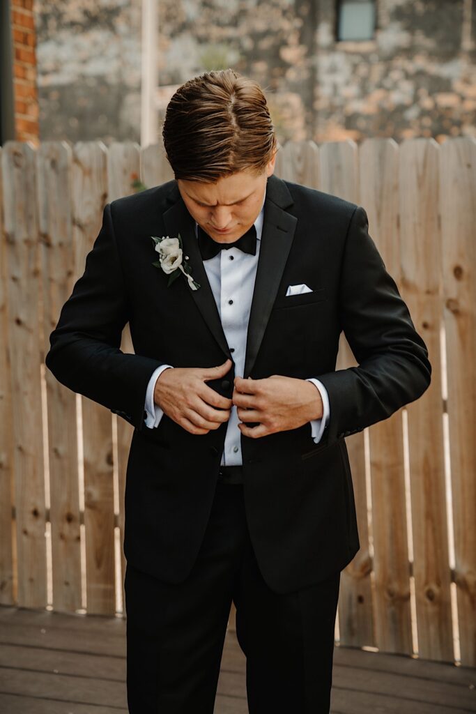Portrait photo of a groom standing outside looking at his suit coat while buttoning it up