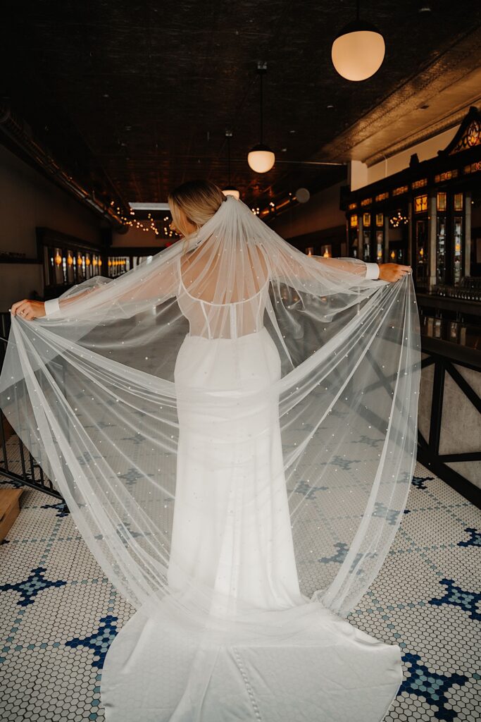 Portrait photo of a bride facing away from the camera and holding her veil out to either side of her with her arms extended in her indoor wedding venue