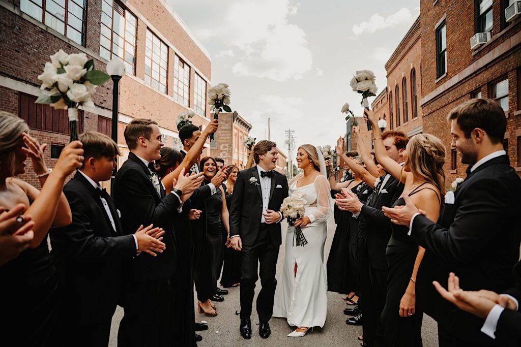 A bride and groom walk with one another while their wedding parties make a tunnel on either side of them cheering them on as they walk down a street in Champaign Illinois after their wedding ceremony at Venue CU