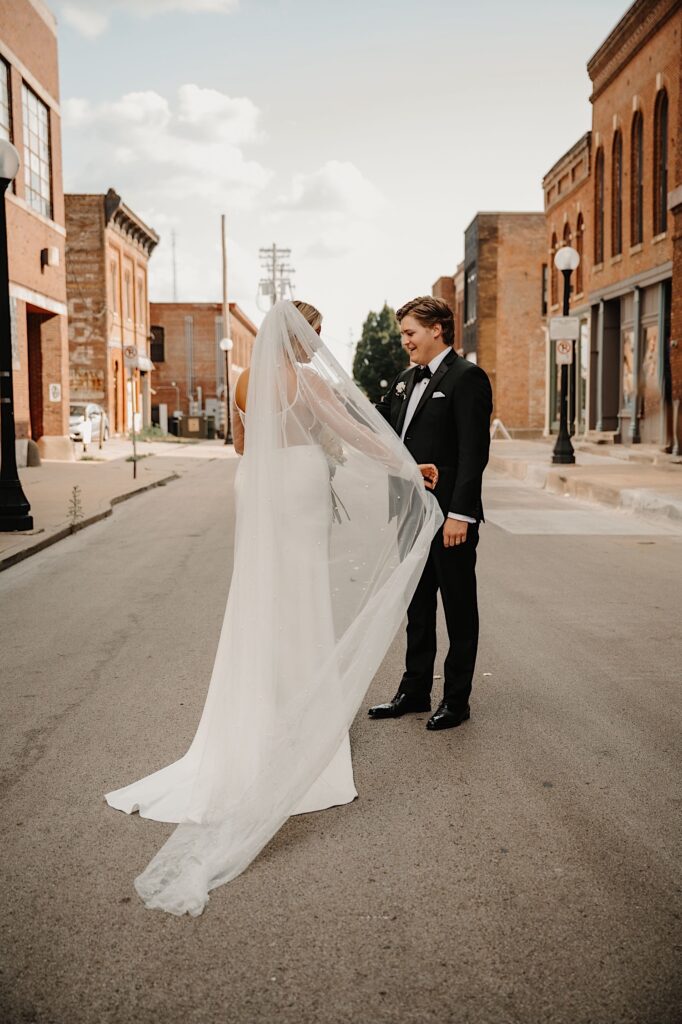 A bride and groom stand on a street in Champaign Illinois as the bride plays with her veil and the groom watches