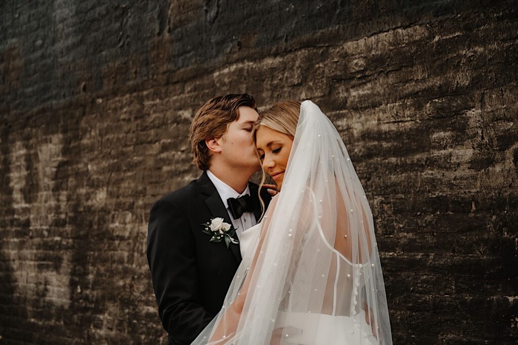 A bride looks down while the groom kisses her on the head as they stand in front of a black brick wall outside their wedding venue, Venue CU in Champaign