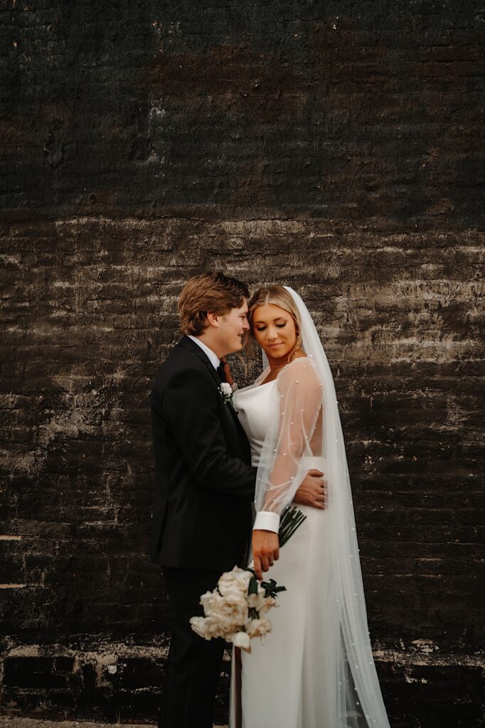 A bride and groom stand together in front of a black brick wall, the bride is looking down at the ground while the groom embraces and looks at her
