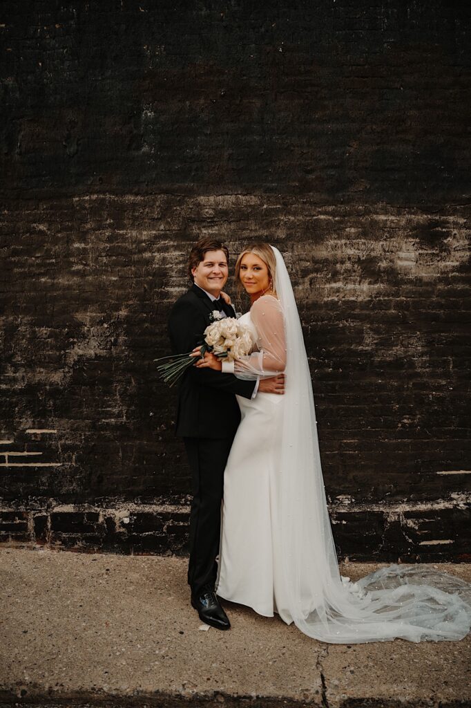 A bride and groom embrace in front of a black brick wall and smile while looking at the camera