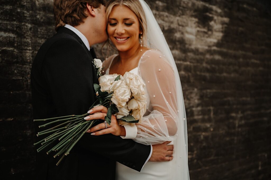 A bride holding her bouquet smiles as the groom kisses her on the cheek as they stand in front of a black brick wall outside of their wedding venue, Venue CU in Champaign