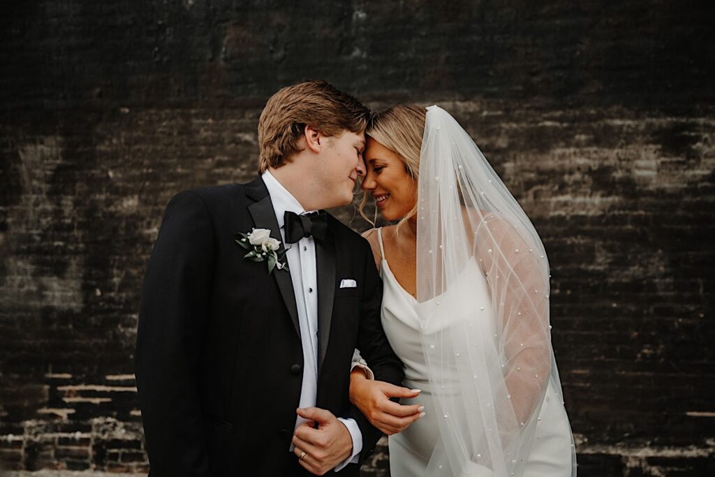 A bride and groom touch foreheads together with their eyes closed and arms interlocked while in front of a black brick wall outside of their wedding venue, Venue CU in Champaign