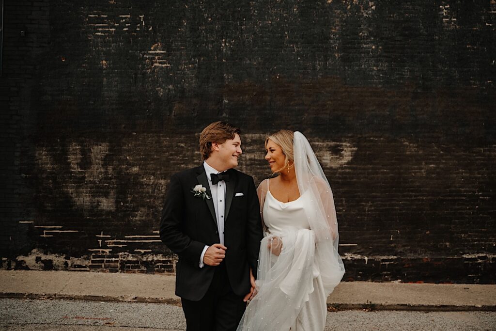 A bride and groom walk hand in hand facing the camera and smile at one another while walking away from a black brick wall outside of their wedding venue, Venue CU in Champaign