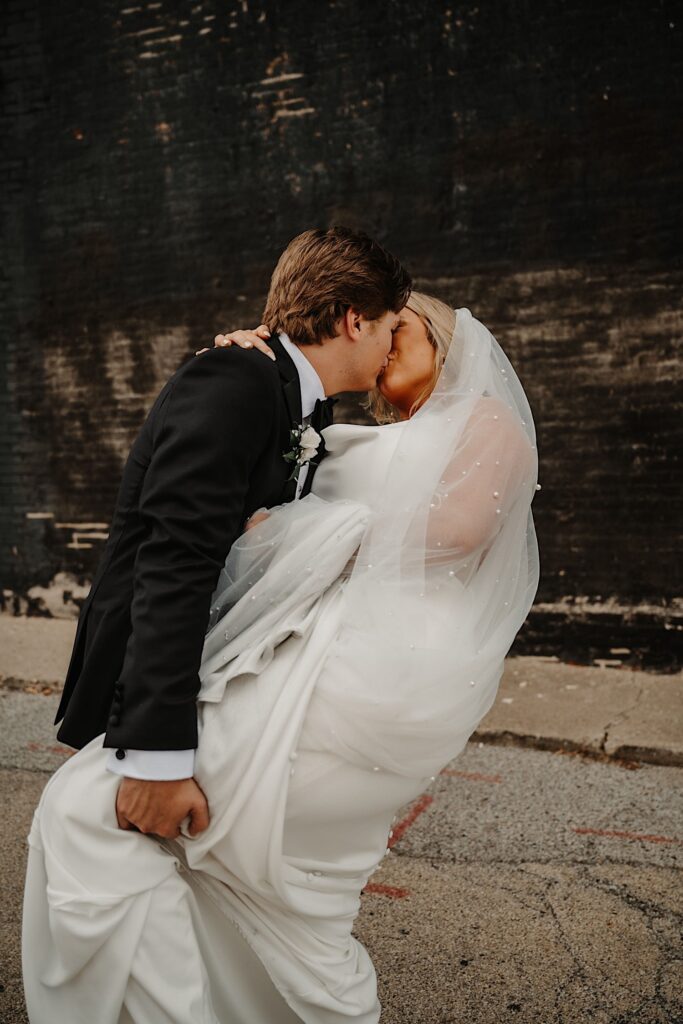 A bride is kissed and dipped by the groom while the two stand outside in front of a black brick wall