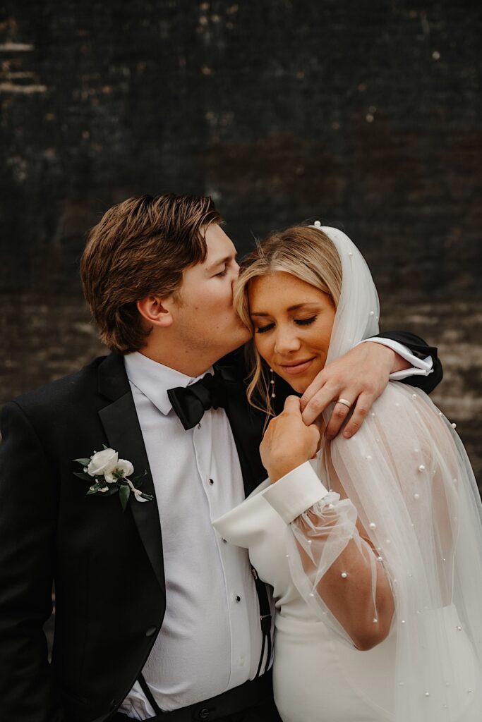 A bride with her eyes closed smiles as the groom kisses her on the head and wraps his arm around her with a black brick wall in the background behind them
