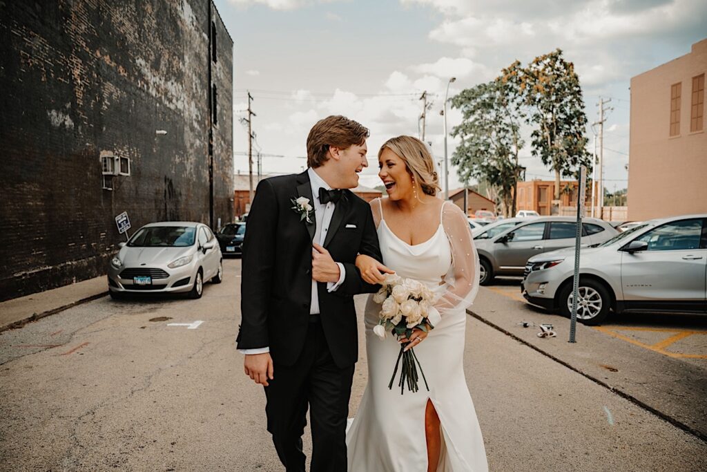 A bride and groom walk together with their arms locked and laugh with one another while outside their wedding venue, Venue CU in Champaign