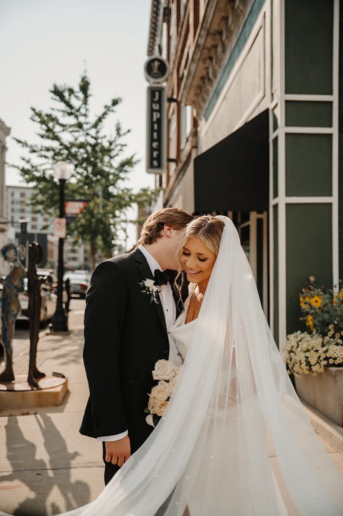 A bride smiles while the groom kisses her on the cheek as the two stand outside on a sidewalk in Champaign Illinois