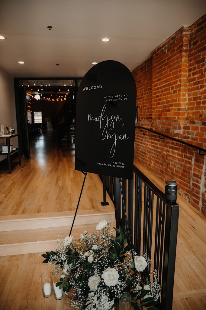 A sign of a wedding reception sits next to a brick wall and a bouquet of flowers