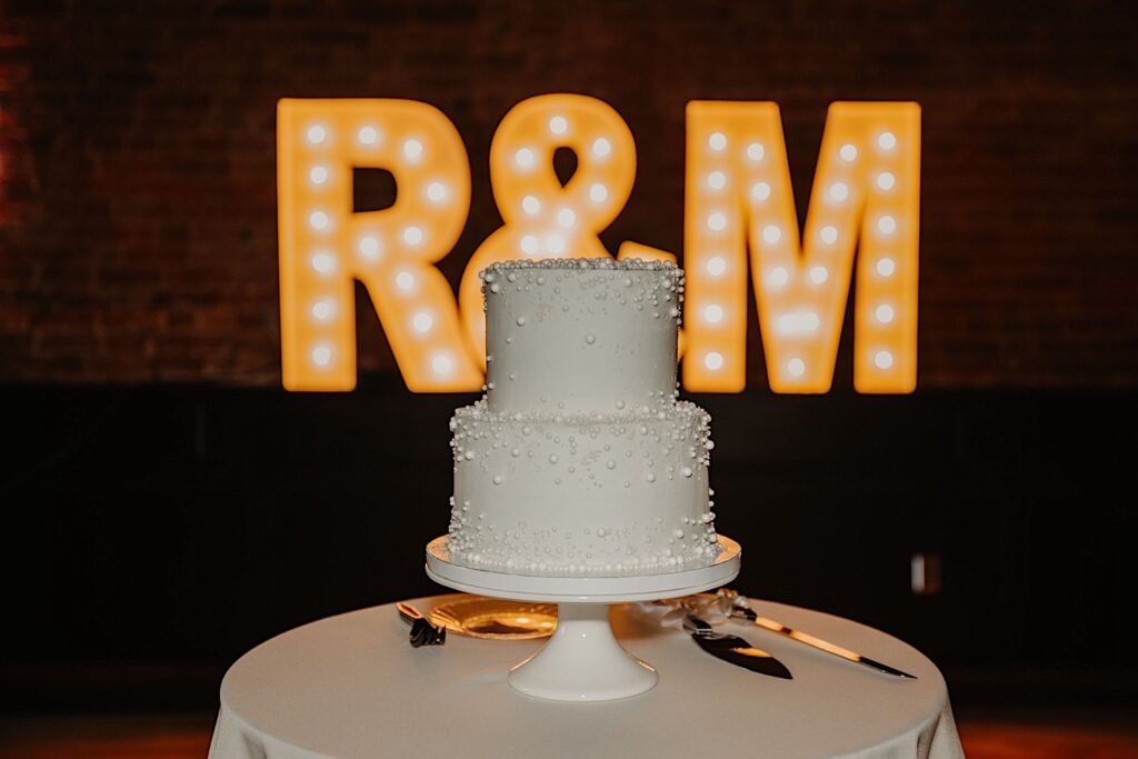 A wedding cake sits on a cake table with large lit up letters on a brick wall behind it that read "R&M" during a wedding reception at Venue CU in Champaign