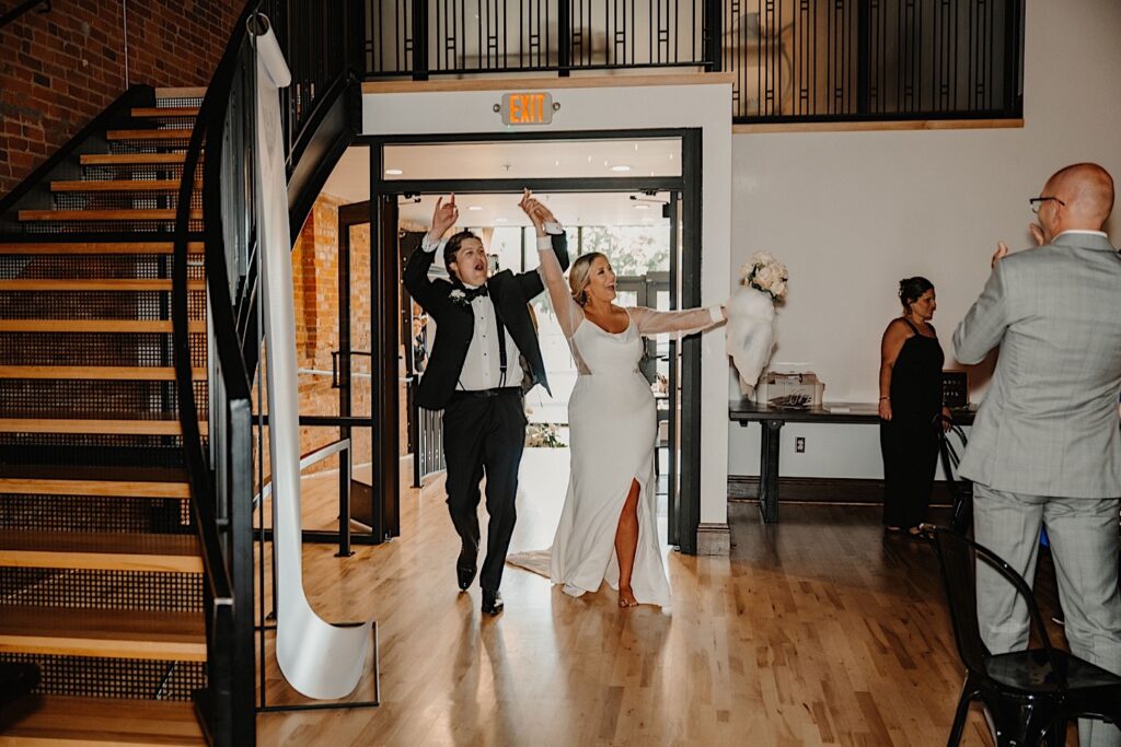 A bride and groom exclaim and raise their arms in the air while entering their wedding reception at Venue CU in Champaign as a few guests in the photo cheer them on
