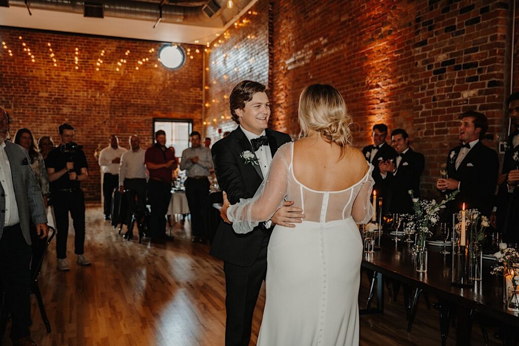 A bride and groom stand in front of their head table while looking at and holding one another as guests of their wedding reception at Venue CU in Champaign all stand around them and cheer