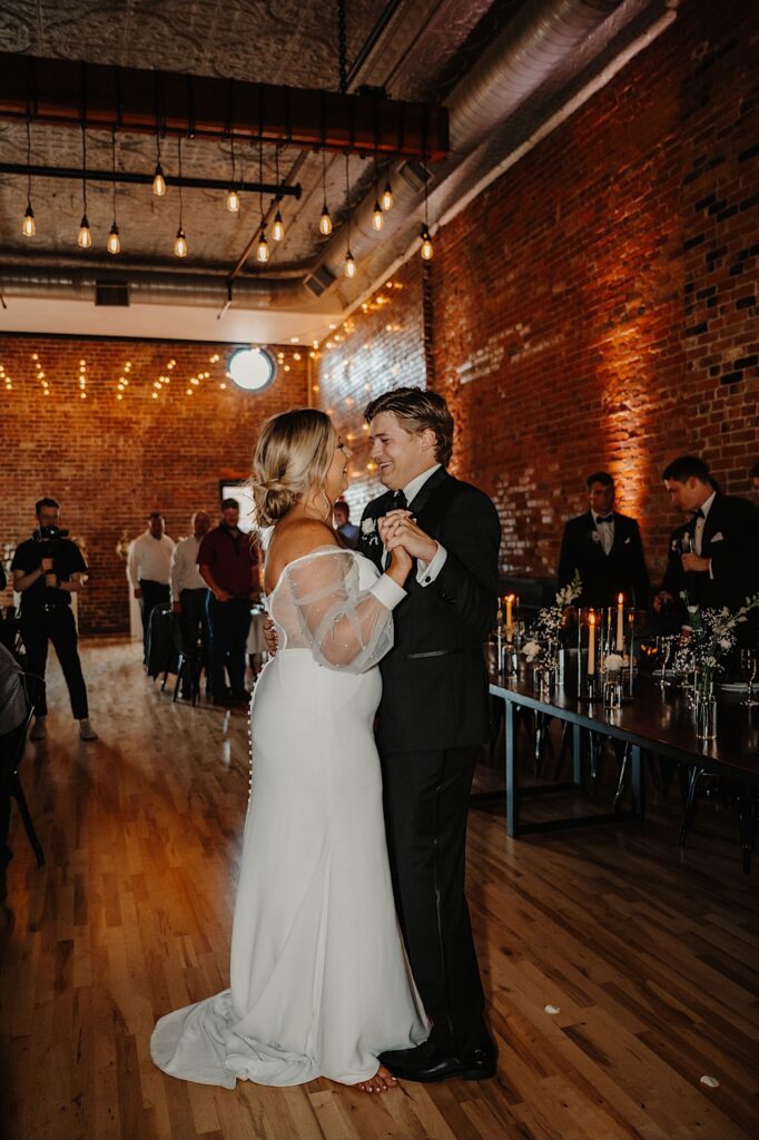 A bride and groom share their first dance during their wedding reception in their brick reception space as guests cheer around them