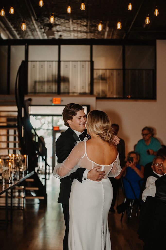 A groom smiles at the bride as the two share their first dance during their indoor reception as guests watch