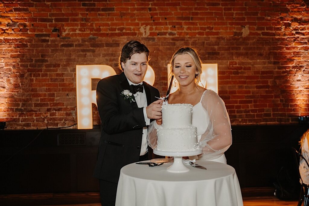 A bride and groom cut their wedding cake during their indoor wedding reception at Venue CU in Champaign, behind them is a brick wall and large electric letters of their initials. 