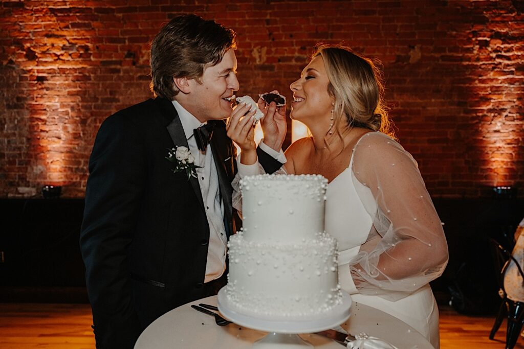 A bride and groom feed one another a piece of their wedding cake while smiling during their wedding reception at Venue CU in Champaign with a brick wall behind them