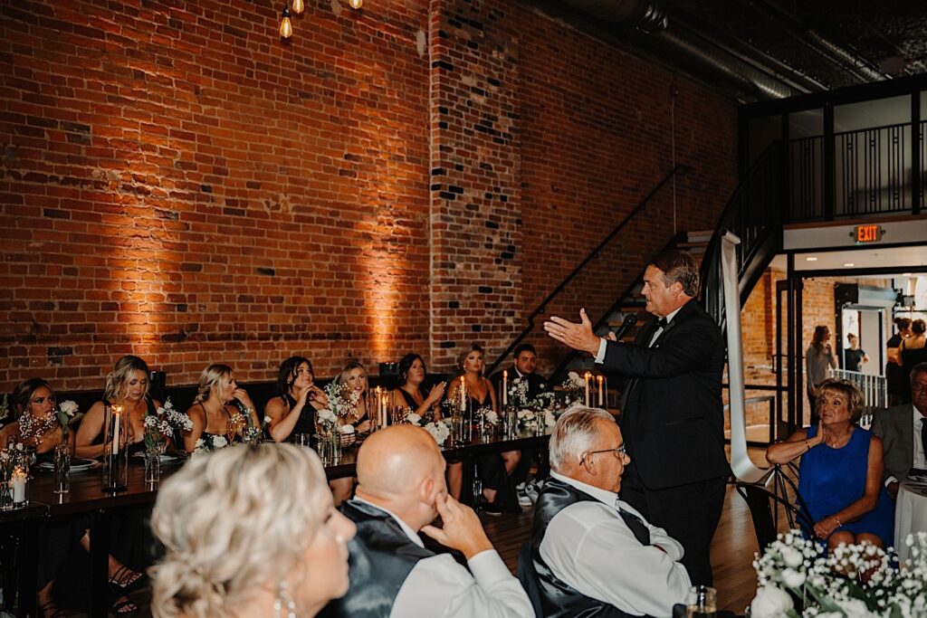 A father stands and gives a speech while gesturing to the head table during a wedding reception inside Venue CU in Champaign as guests watch