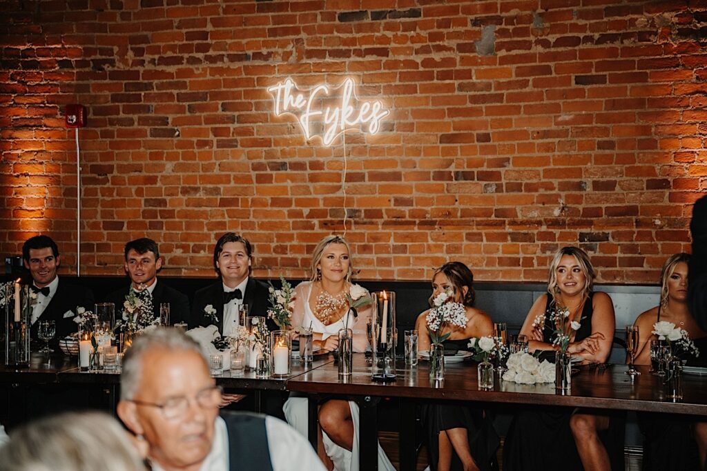 A bride and groom sit at their head table and smile next to their wedding parties as speeches are given, behind them is a brick wall and a neon sign of their last name hangs above them, they are in the indoor reception space of their venue, Venue CU in Champaign