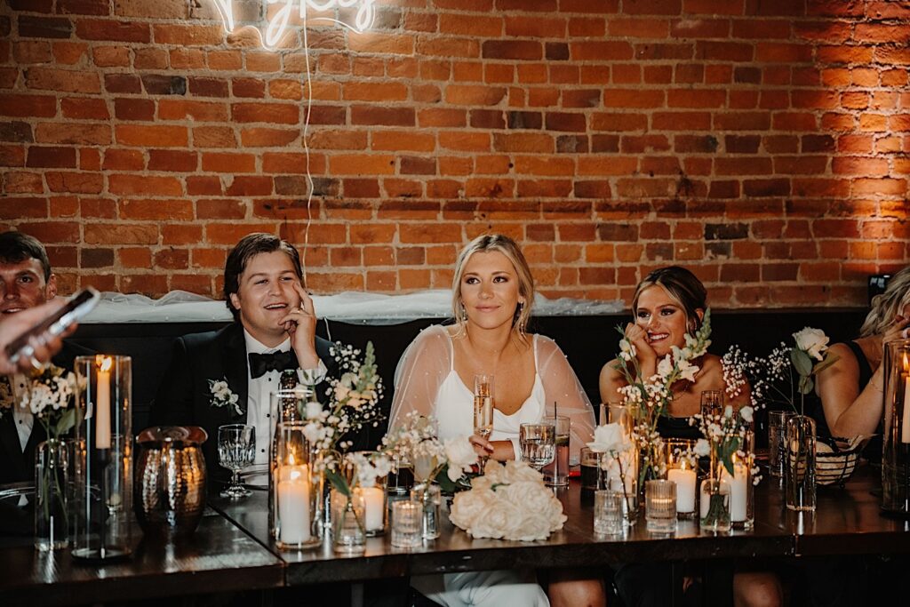 A bride and groom smile while sitting at their head table next to their wedding parties during their indoor reception at Venue CU in Champaign
