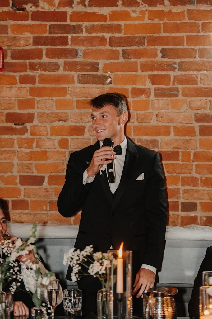 A groomsmen stands in front of a brick wall with a microphone to give a speech during a wedding reception