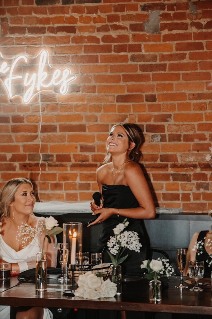 A bridesmaid laughs while standing next to the bride who is seated at her head table, the bridesmaid is about to give a speech during the indoor wedding reception 