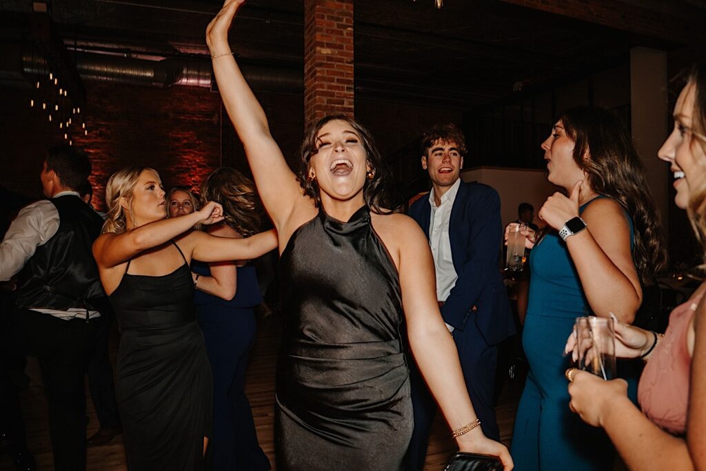 Guests of a wedding reception at  Venue CU in Champaign Illinois dance on the dancefloor together, one guest is gesturing towards the camera while smiling