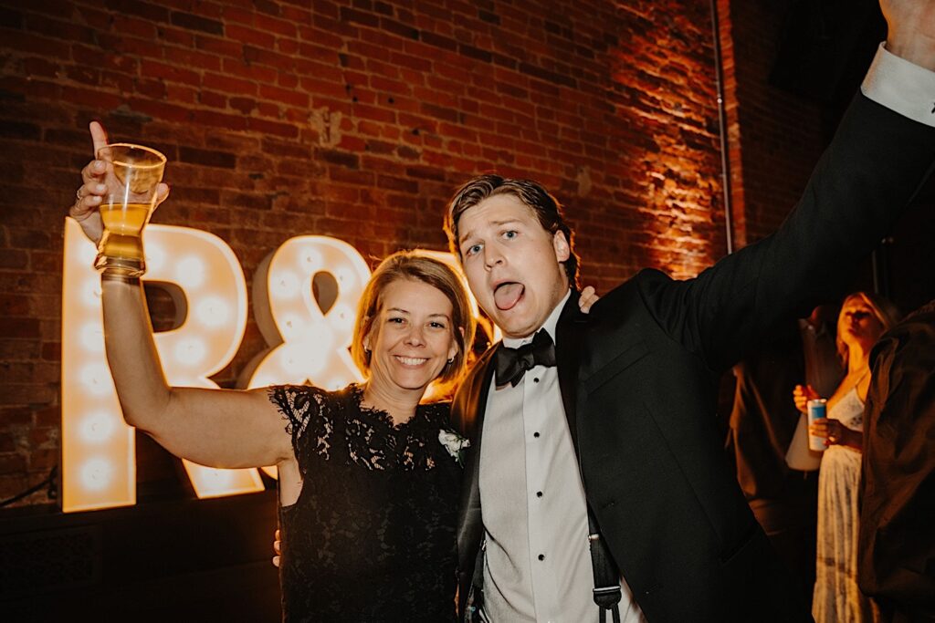 A groom and a guest of his wedding reception at Venue CU in Champaign Illinois gesture and smile towards the camera with a brick wall and lit up letters in the background