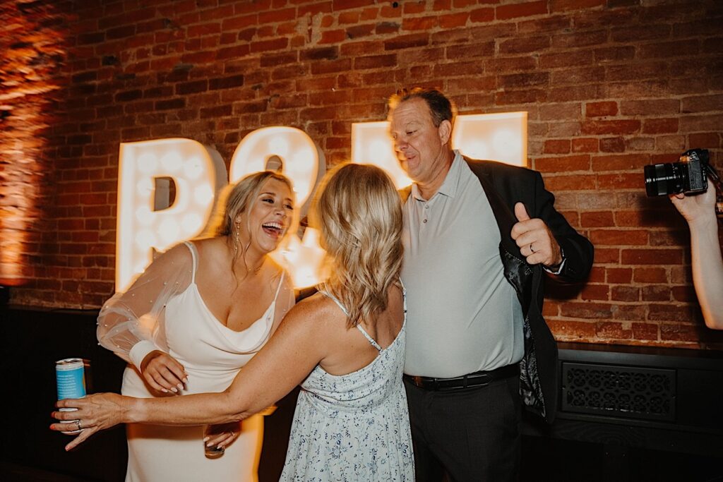 A bride smiles while talking to guests of her wedding reception at Venue CU in Champaign Illinois with a brick wall and large lit up letters behind them